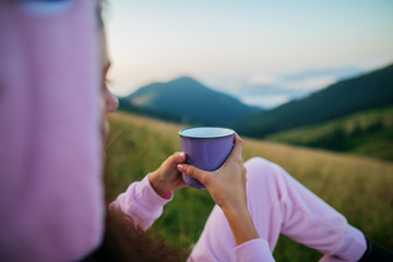 Closeup photo of cup with coffee in traveler's hand over out of focus mountains view. Young woman in a pink dragon costume drinks a hot drink from a cup and enjoys the scenery in the mountains