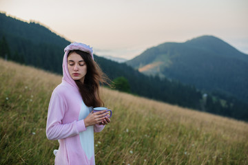Young woman in pink dragon costume drinking coffee on top of mountain. Incredible mountain landscape at sunrise. Calm and inspiration on high peak of mountain
