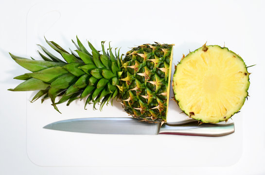 An image of a half-cut pineapple with a steel-blue knife nearby on a white breadboard on a white background