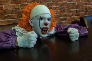 A teenager in a scary clown costume with sharp teeth sits at a cafe table and clenched his hands...