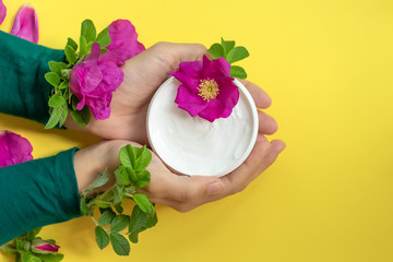 Obraz na płótnie Canvas Young female hands are holding jar with white anti-ageing moisturizing cream with dog rose oil essential and vitamin E on bright yellow background with bright pink dog roses, petals and green leaves