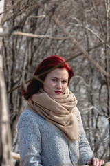Pretty young women with red hair in beautiful beige scarf and grey coat posing in a winter forest or park.