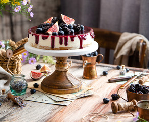 homemade tasty whole cheesecake decorated with figs, blackberries, blueberries and purple sauce on top served on wooden cake stand on grey table with flowers and berries, selective focus