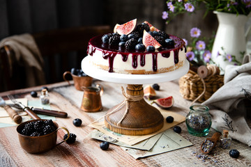 homemade tasty whole cheesecake decorated with figs, blackberries, blueberries and purple sauce on top served on wooden cake stand on grey table with flowers and berries, selective focus
