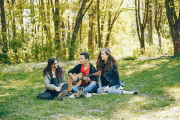 Naklejka na ściany i meble Three friends sitting on a green grass. Boy playing on a guitar with his two girlfriends. Two girls have fun with their friend