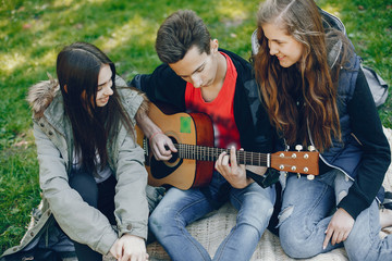 Obraz na płótnie Canvas Three friends sitting on a green grass. Boy playing on a guitar with his two girlfriends. Two girls have fun with their friend
