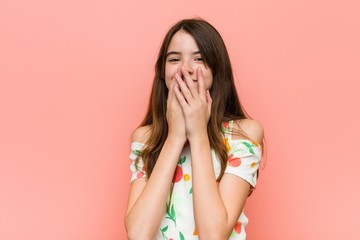 Girl wearing a summer clothes against a red wall laughing about something, covering mouth with hands.