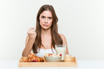 Obraz na płótnie Canvas Young caucasian woman having breakfast showing fist to camera, aggressive facial expression.
