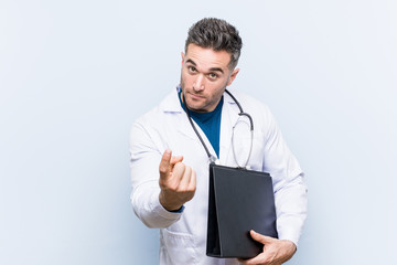 Caucasian doctor man holding a folder pointing with finger at you as if inviting come closer.