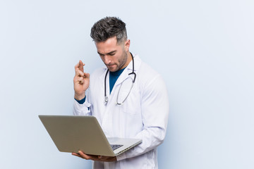 Caucasian doctor man holding a laptop crossing fingers for having luck