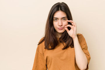 Young brunette woman against a beige background with fingers on lips keeping a secret.