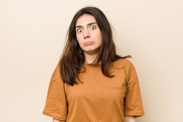 Young brunette woman against a beige background shrugs shoulders and open eyes confused.