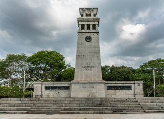 Obraz premium Singapore - March 20, 2019: Gray stone Cenotaph war memorial surrounded by green foliage in Esplanade Park under heavy gray sky promising rain. World War One side.