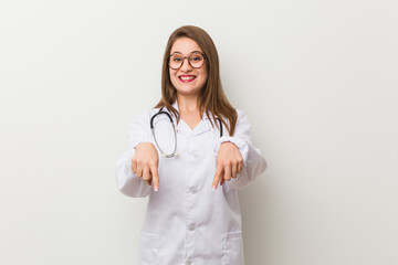 Young doctor woman against a white wall points down with fingers, positive feeling.