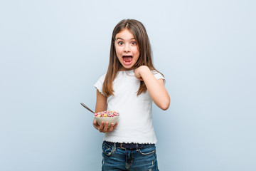 Little caucasian girl holding a cereal bowl surprised pointing at himself, smiling broadly.