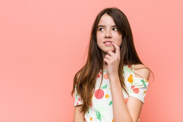 Girl wearing a summer clothes against a red wall looking sideways with doubtful and skeptical expression.