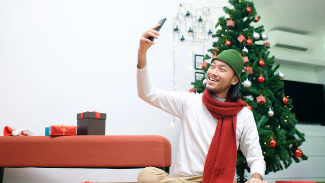 Young asian man take selfie, video call by smart phone while sitting in Christmas celebration party at home with happiness