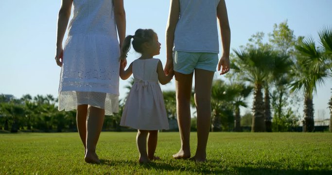 little girl is holding hands of her older sister and mother and walking in sunny day in park, back view