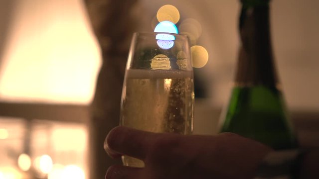 Male hand holds green bottle of cold sparkling champagne and fills a transparent glass with a drink on golden bokeh background, close-up. Romantic dinner in intimate atmosphere under muffled lighting.