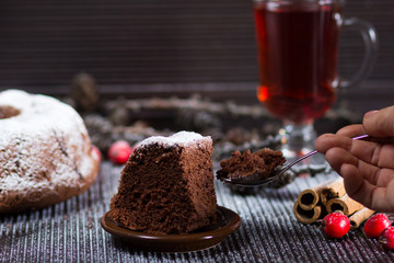 Piece of christmas chocolate sponge cake with sugar powder on wooden table with glass of red tea and sticks of cinnamon