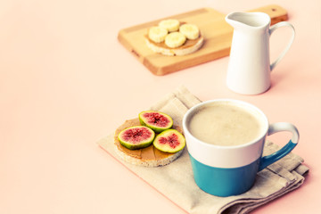 Healthy breakfast: coffee latte, non dairy milk, sandwiches with banana and figs on a cutting board, pink background.