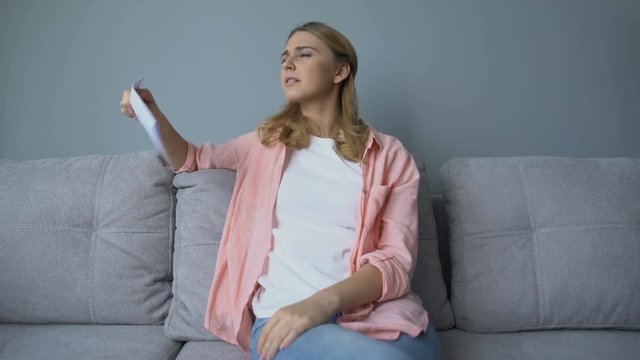 Girl waving paper sitting on couch, suffering heat, needs air conditioner