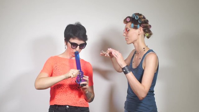 two girls smoke cannabis, one smokes bong, the second smokes a joint