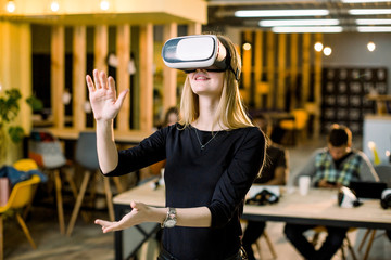 Cheerful Caucasian business woman wearing virtual reality glasses in office. Young girl in casual wear having fun with vr goggles while her coworkers sitting at the table