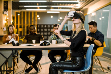 Business team of multiethnical people working on virtual reality applications and games, young excited woman testing VR glasses or goggles sitting in the office room with coworkers