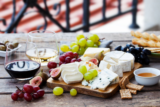 Cheese and fruits assortment on cutting board with red, white wine on wooden background. Copy space.