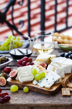 Cheese and fruits assortment on cutting board with red and white wine on wooden background. Copy space.