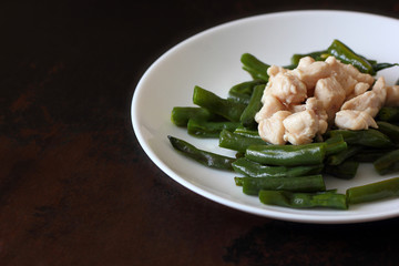 Baked string beans and pieces of chicken breast in a white plate on a wooden countertop.