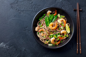 Stir fry noodles with vegetables and shrimps in black bowl. Slate background. Top view. Copy space.