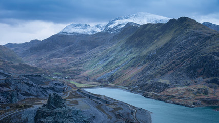 Beautiful landscape image of Dinorwig Slate Mine and snowcapped Snowdon mountain in background during Winter in Snowdonia with Llyn Peris in foreground