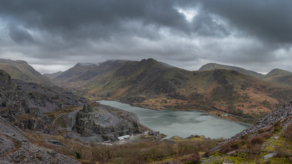 Beautiful landscape image of Dinorwig Slate Mine and snowcapped Snowdon mountain in background during Winter in Snowdonia with Llyn Peris in foreground