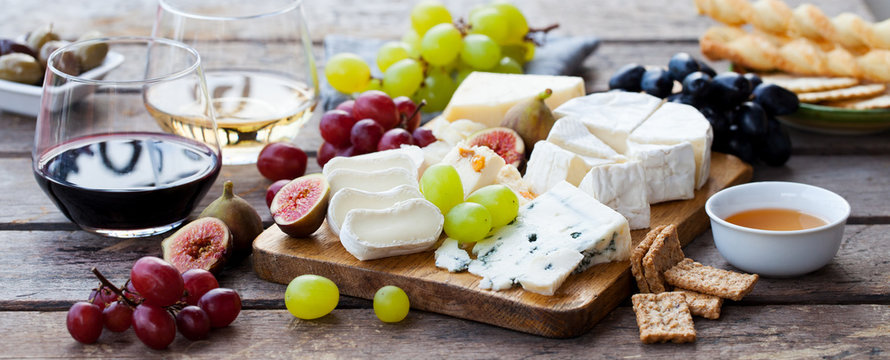 Cheese and fruits assortment on cutting board with red, white wine on wooden background.
