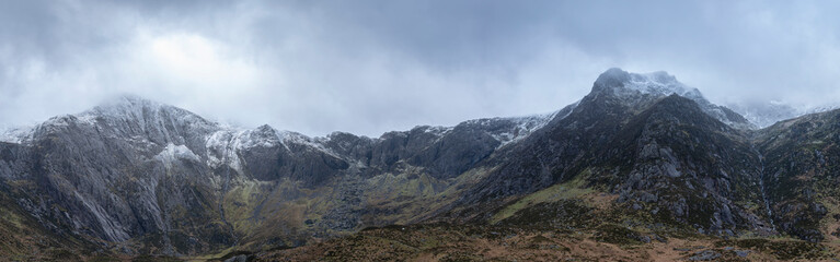 Fototapeta na wymiar Stunning dramatic panoramic landscape image of snowcapped Glyders mountain range in Snowdonia during Winter with menacing low cloudshanging at the mountain peaks