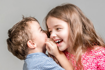 Boy in a blue shirt whispers in the ear a girl in a red dress on a gray background.