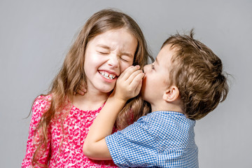 Boy in a blue shirt whispers in the ear a girl in a red dress on a gray background. Isolated.