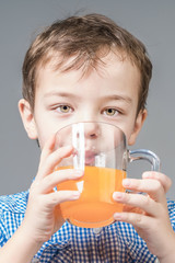 Cute boy in a blue shirt drinks orange juice on a gray background.