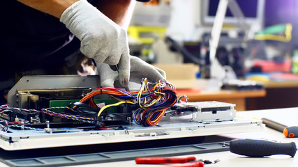 Technician works in service center. Repairman detaches power supply repairs computer monoblock in workshop. He disconnects wires, takes out and puts detail on table, continues to diagnose monitor.