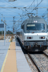 A grey and white train with wire fences and blue sky with white background clouds.