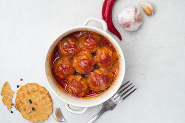 Meatballs stewed in tomato sauce in a saucepan, pepper and garlic on white table