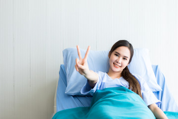 Asian young female patient smiley face lifts two fingers up fighting with illness on bed in the room hospital background.