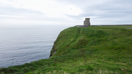 Photograph of an old lighthouse in front of the Cliff of Moher in Ireland; behind the very green lawn.