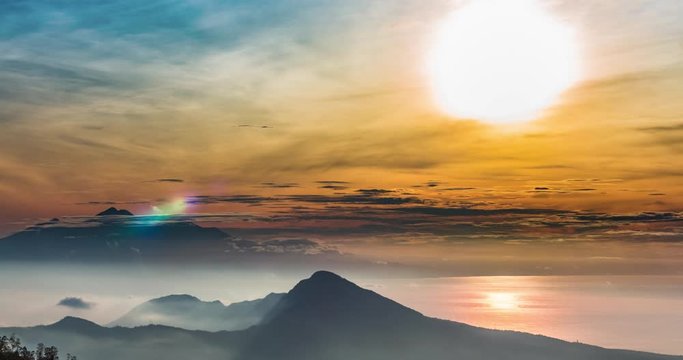 Time lapse of sunrise above mountain valley in mist and Rinjani volcano in afar. Warm changing colors and floating clouds above the mountains, view from slope of Agung volcano.