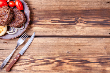 Fresh grilled beefsteaks with vegetables - onions and tomatoes - on brown vintage style plate on wooden background with fork and knife. Grilled food. Copy space.