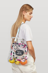 Three quarter half-turn shot of a blond girl, wearing white t-shirt and ivory trousers. She has beach clear bag with cosmetics print and lettering "wake up and makeup" on her shoulder.