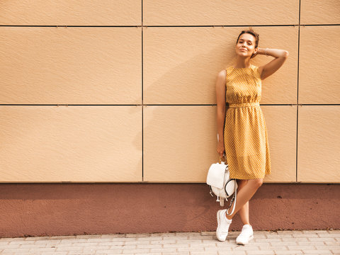 Portrait of beautiful smiling hipster model dressed in summer yellow dress. Trendy girl posing in the street background. Funny and positive woman having fun