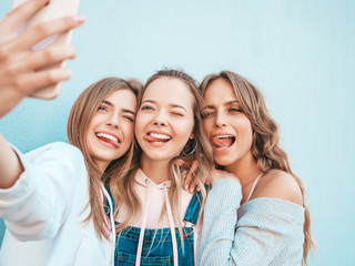 Fototapeta premium Three young smiling hipster women in summer clothes.Girls taking selfie self portrait photos on smartphone.Models posing in the street near wall.Female showing positive face emotions